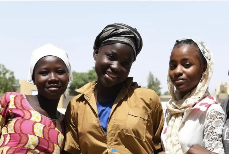 There adolescent girls wearing a variatey of head covers look at the camera against a light blue sky