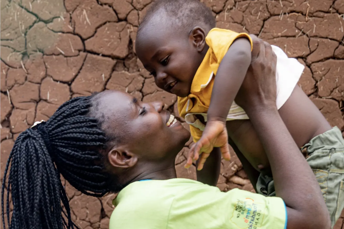 A smiling mother wearing a green t-shirt holds her infant up to her face